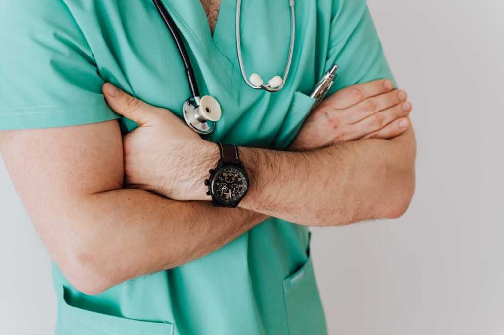 pexels-photo-4021769 Close-up of a male doctor in scrubs with crossed arms and stethoscope on white background.